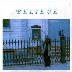 FRØKEDAL - Believe; Foto von eine Frau, die vor einem eingezäunten Haus auf dem Gehweg steht