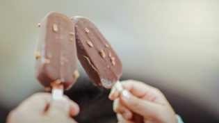 zwei Hände, die ein Eis am Stiel mit Schokolade überzogen halten und damit anstoßen