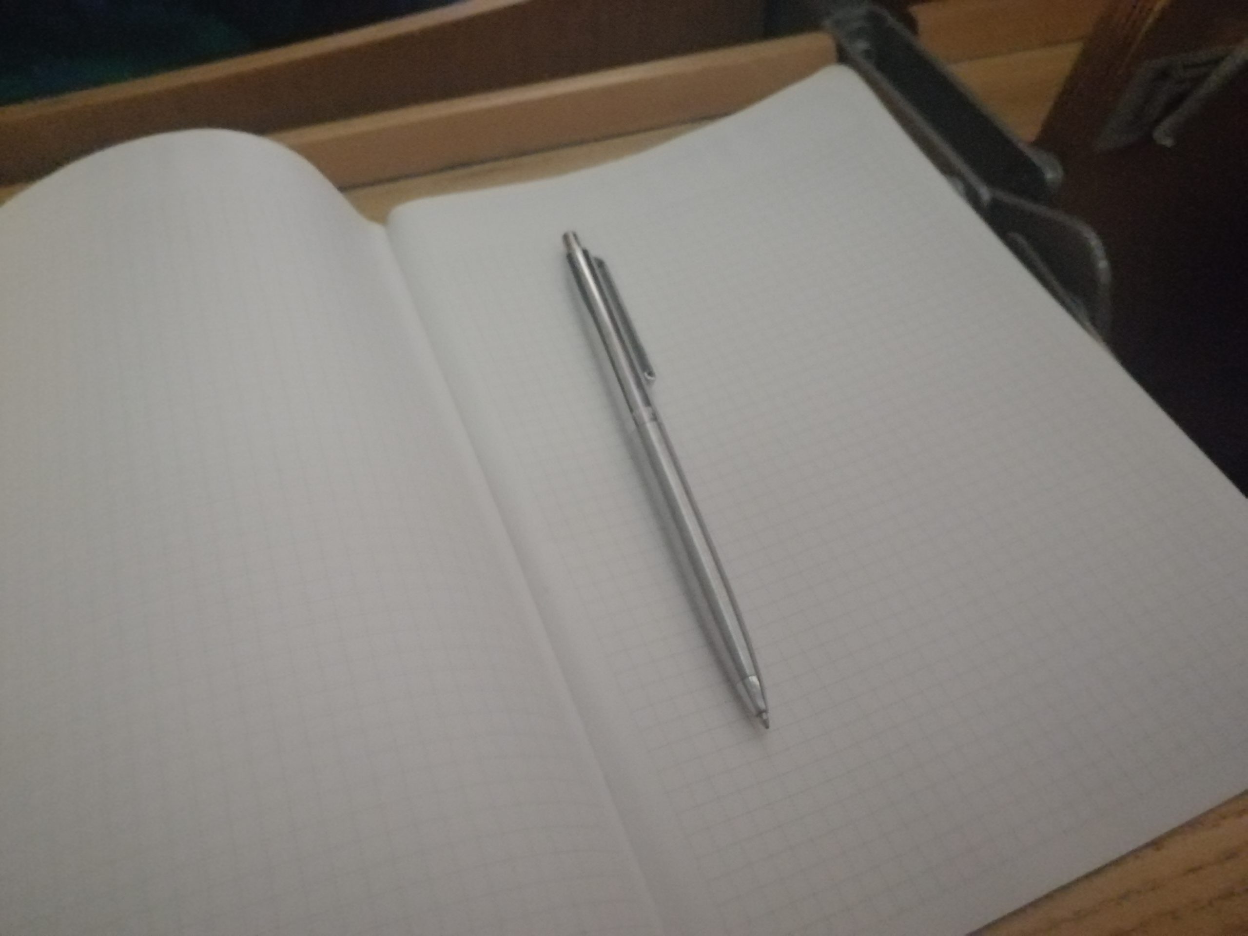 Weißes Schulheft mit kariertem Papier auf dem ein silbernen Kugelschreiber liegt.