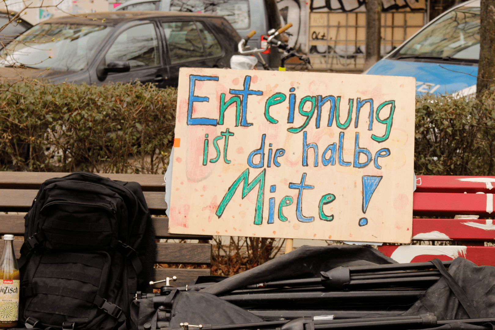 Eine Bank vor einer Hecke, auf der ein Rucksack und eine Tasche liegen; auf einem Pappschild steht in bunter Handschrift: "Enteignung ist die halbe Miete!"